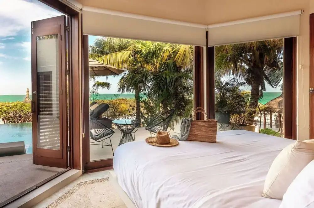 Blick aus dem Hotelzimmer über das Bett auf eine Terrasse mit Palmen und dem Meer.