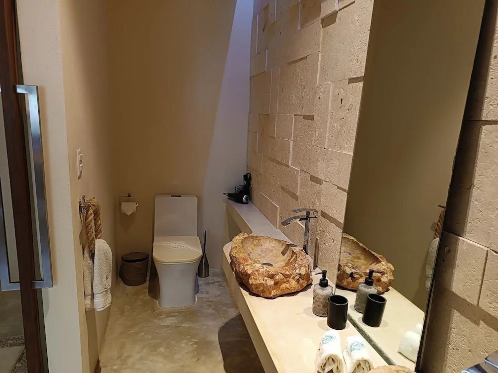 Badezimmer mit Natursteinwaschbecken, großem Spiegel und Toilette vor beigefarbener Wand.