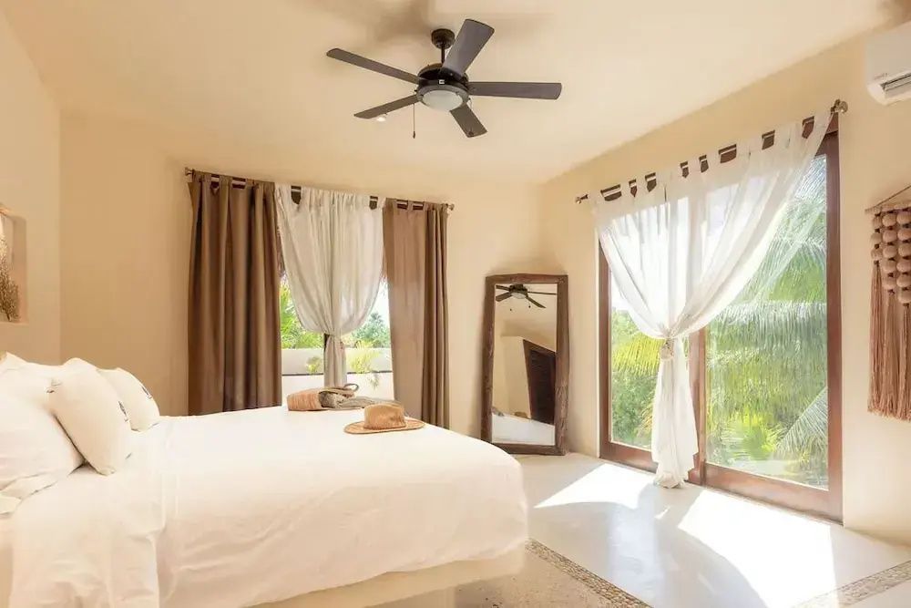 Schlafzimmer mit großem Doppelbett, Vorhängen an den Glastüren, Deckenventilator und Holzspiegel.