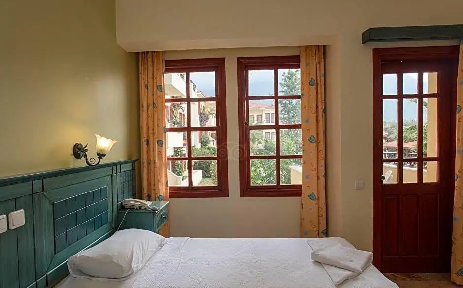 Doppelbett mit weißer Bettwäsche, grünem Kopfteil und Blick durch zwei Holzfenster ins Grüne.