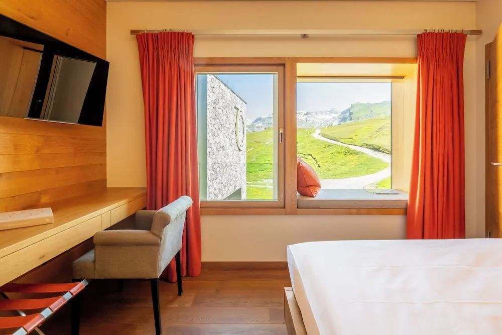 Hotelzimmer mit Schreibtisch, rotem Vorhang und Fensterplatz mit Blick auf die Alpen.
