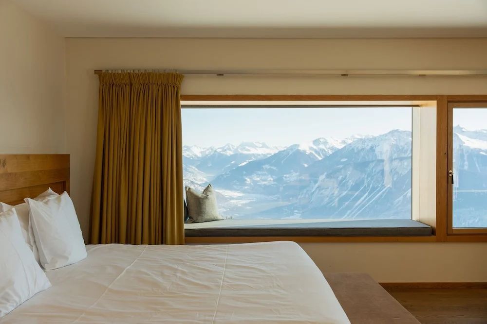Blick durch ein großes Fenster im Hotelzimmer auf verschneite Berge und das Tal.