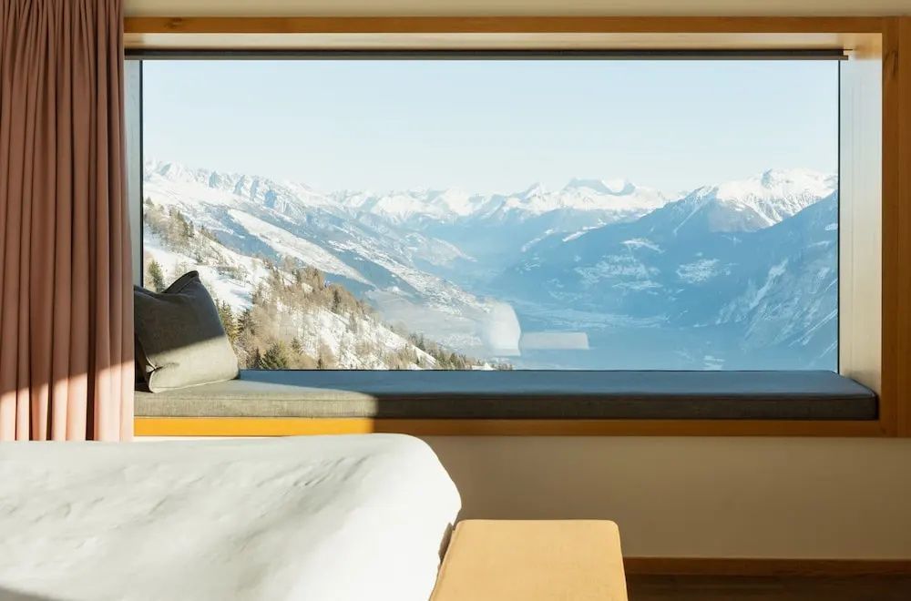 Großes Panoramafenster mit Sitzbank und Blick auf sonnige verschneite Berggipfel.