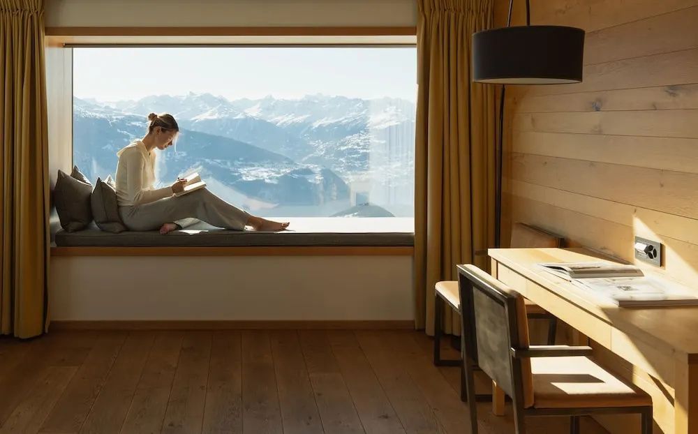 Frau sitzt in der Fensternische eines Holzzimmers und blickt auf die Berge.