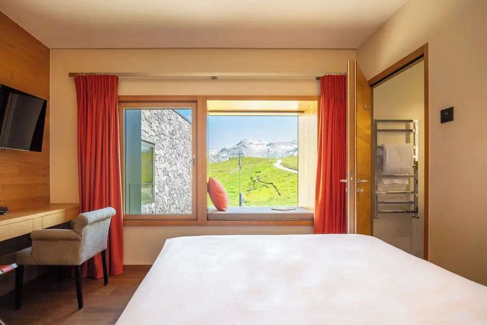Schlafbereich im Hotelzimmer mit großem Fenster, Sitznische und Ausblick auf die Berge.