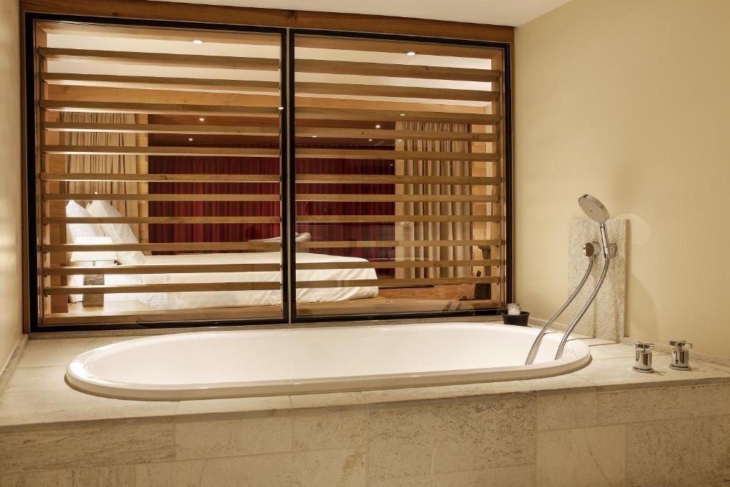 Ovale Badewanne im Badezimmer mit Fenster aus Holzlamellen zum angrenzenden Schlafbereich.