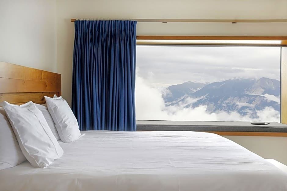 Hotelbett mit weißer Bettwäsche und Vorhang vor Panoramafenster mit Bergblick.
