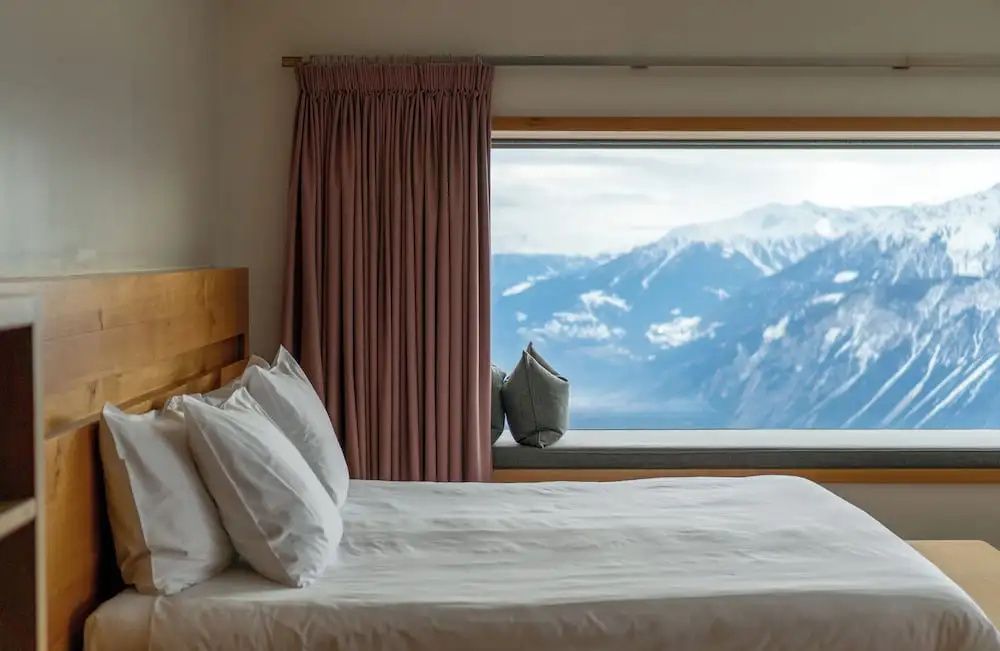 Doppelbett mit weißer Bettwäsche vor einem Fenster mit Bergpanorama.