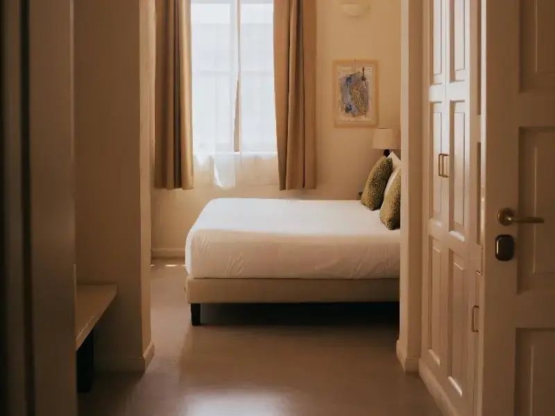 Blick in ein Schlafzimmer mit Doppelbett am Fenster und beigefarbenen Vorhängen.