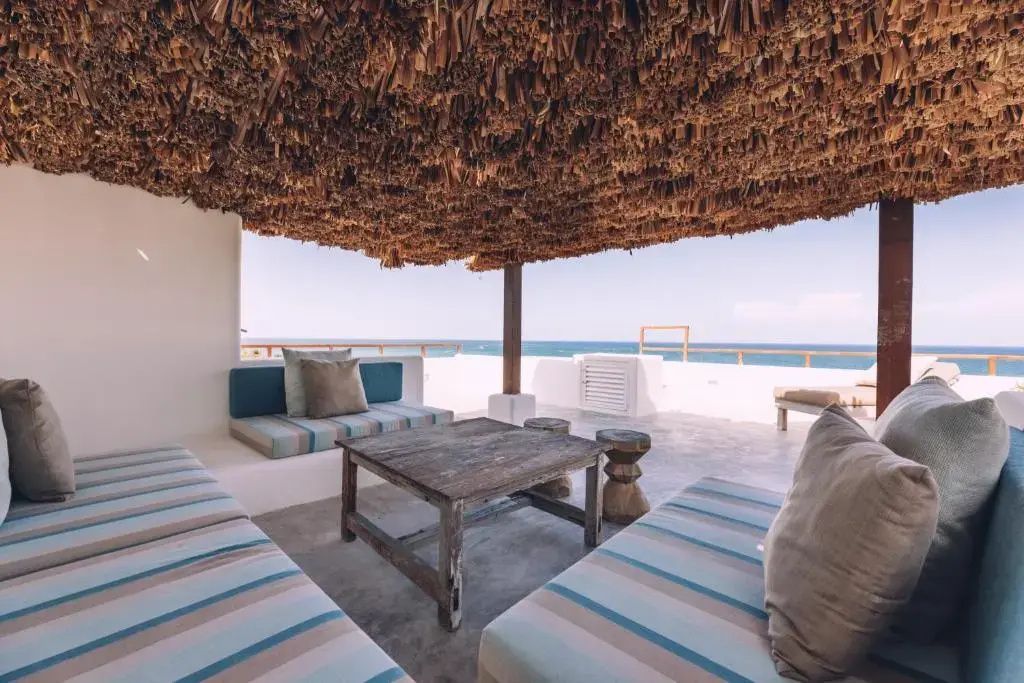Überdachte Terrasse mit Strohdach, gepolsterten Sitzbänken und Blick auf das Meer.