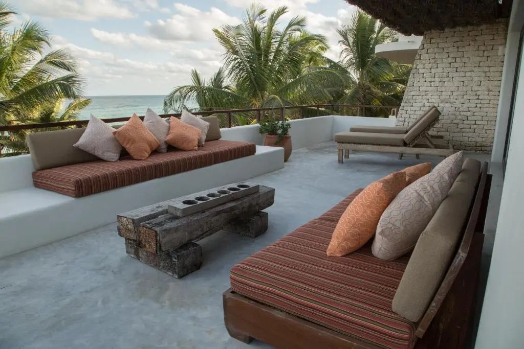 Terrasse mit gepolsterten Sitzbänken, einem Holztisch und Blick auf das Meer und Palmen.