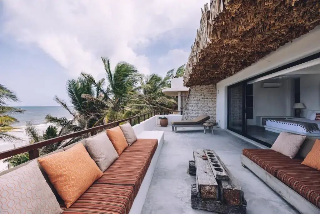 Große Terrasse unter Reetdach mit orangefarbenen Kissen, Liegestühlen und Meerblick.