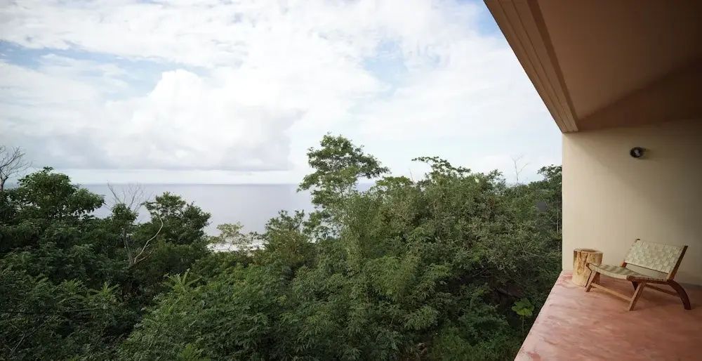 Blick von der Terrasse über grüne Baumkronen auf den Ozean unter bewölktem Himmel.