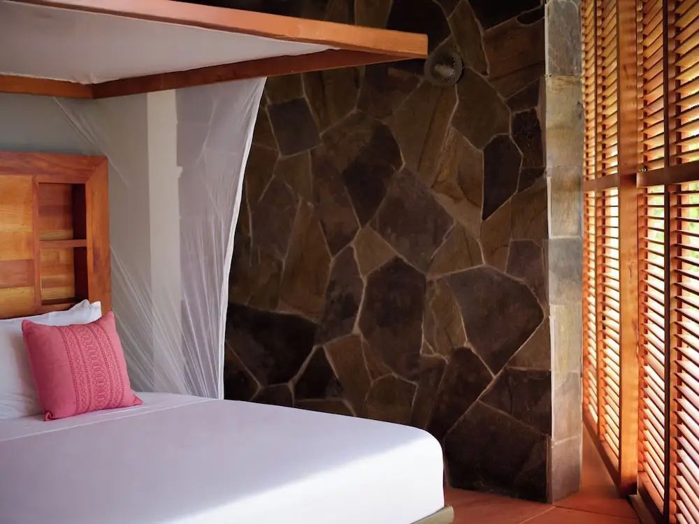Bett mit weißem Moskitonetz und rosa Kissen neben einer dunklen Natursteinwand.