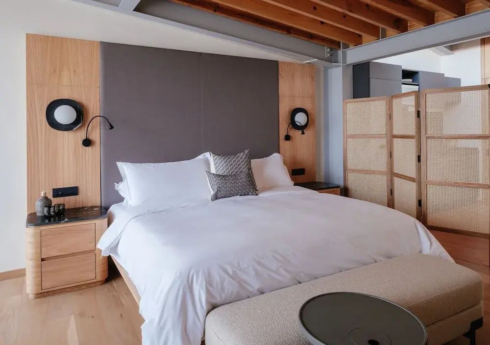 Doppelbett mit weißer Bettwäsche, grauem Kopfteil und Holzdecke in einem Hotelzimmer.