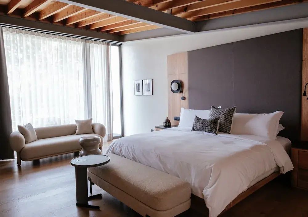 Schlafbereich mit großem Bett, Beistellsofa vor dem Fenster und Holzgebälk an der Decke.