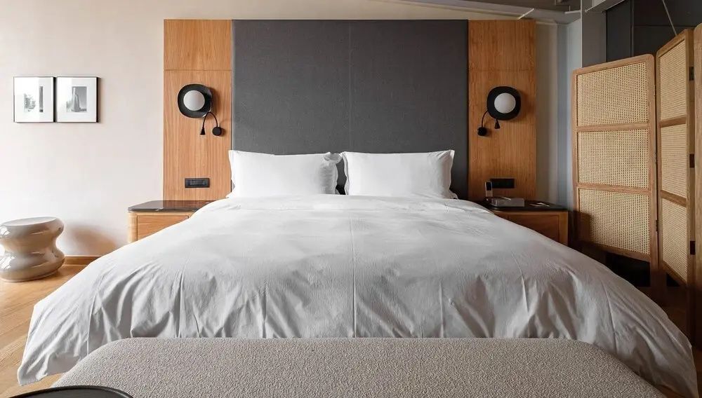 Doppelbett mit weißer Bettwäsche, hölzernem Kopfteil und runden Wandlampen.