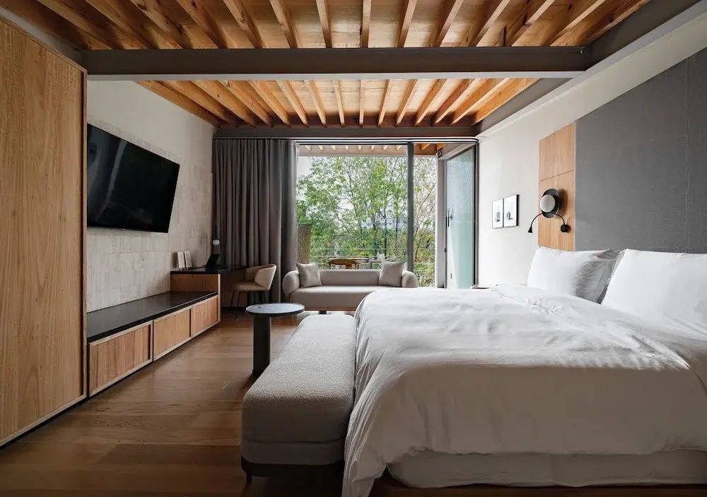 Schlafzimmer mit großem Bett, Fernseher an der Wand und bodentiefen Fenstern ins Grüne.
