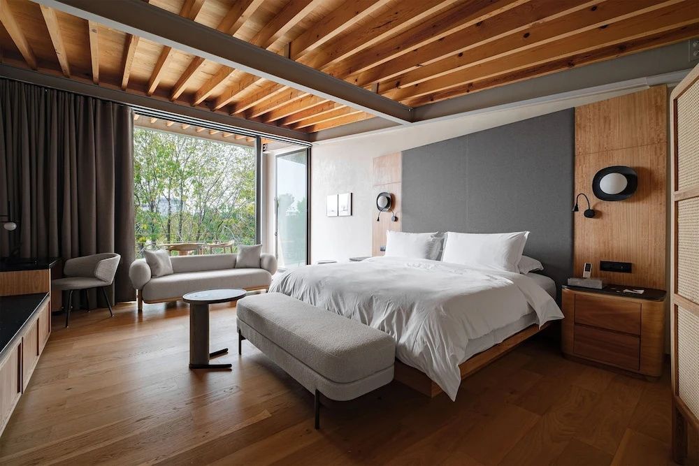 Schlafzimmer mit Holzbalkendecke, weißem Doppelbett und großer Fensterfront.