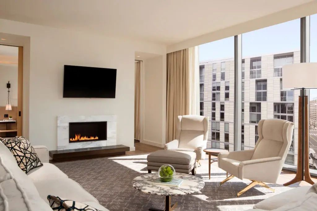 Wohnbereich mit modernem Kamin, Fernseher, hellen Sesseln und bodentiefen Fenstern.