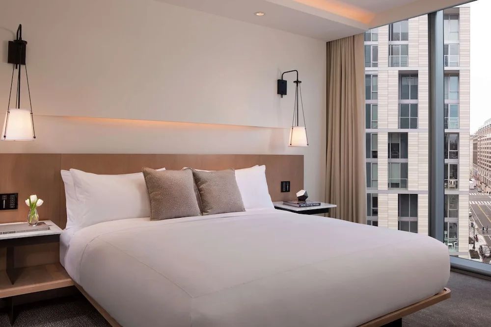 Doppelbett mit weißer Bettwäsche, Nachttischlampe und großem Fenster im Hotelzimmer.