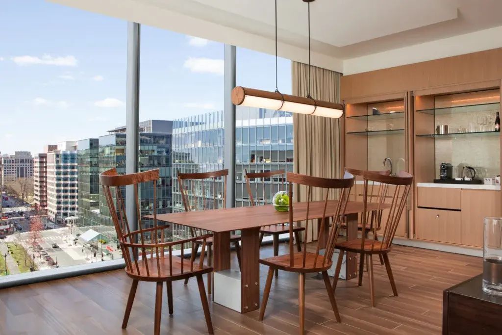 Esstisch aus Holz mit sechs Stühlen vor bodentiefen Fenstern mit Blick auf Stadtgebäude.