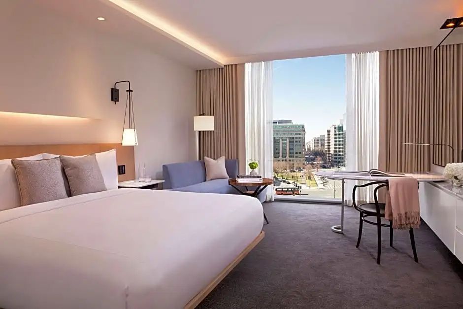 Helles Hotelzimmer mit Doppelbett, blauer Sitzcouch und bodentiefem Fenster mit Blick auf die Stadt.