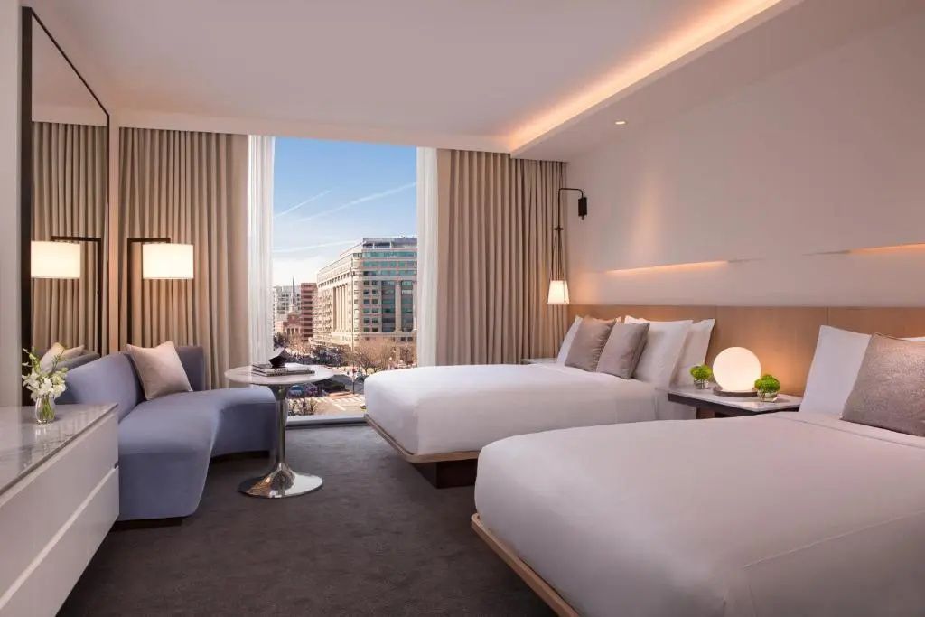 Hotelzimmer mit zwei Doppelbetten, geschwungenem blauen Sofa und Fenster mit Blick auf die Stadt.