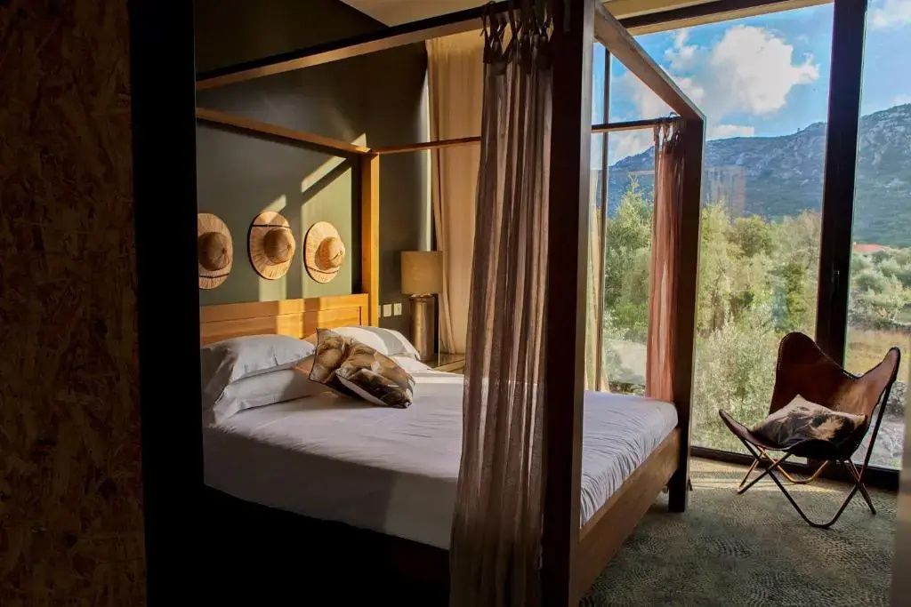 Hölzernes Himmelbett mit weißer Bettwäsche und Vorhängen in einem lichtdurchfluteten Hotelzimmer mit Blick ins Grüne.