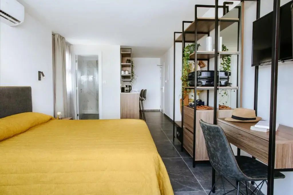 Hell eingerichtetes Hotelzimmer mit gelbem Bett, offenem Regalsystem und Schreibtisch.