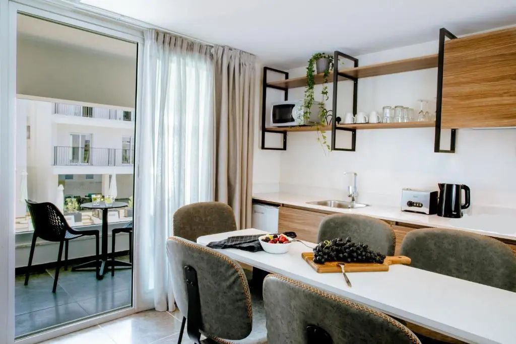 Essenstisch mit Stühlen und Küchenzeile in einem hellen Hotelzimmer mit Zugang zum Balkon.