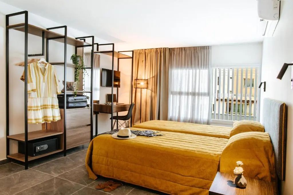Modern eingerichtetes Hotelzimmer mit zwei Betten in Gelb, Holzschreibtisch und offenem Schrank.