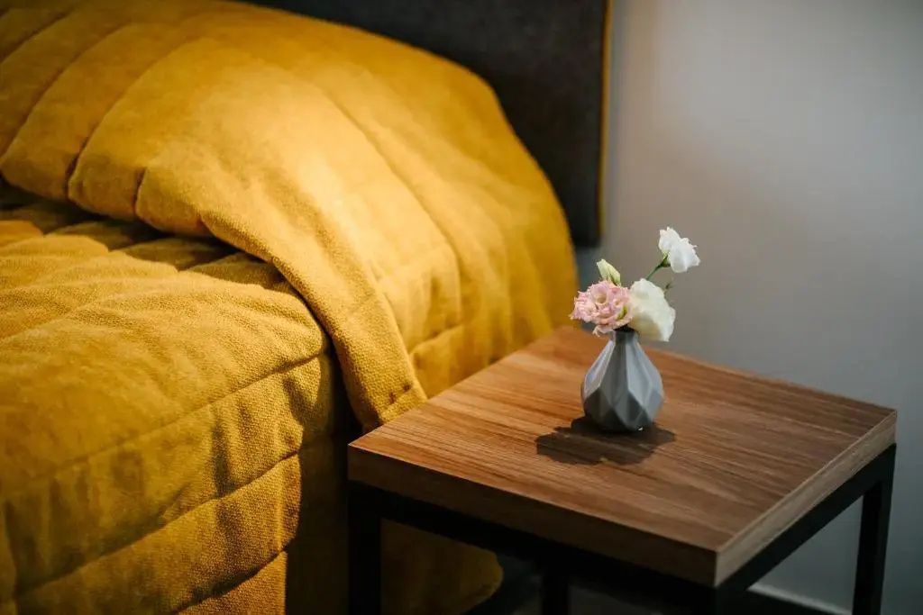 Holznachttisch neben einem Bett mit gelber Tagesdecke und einer kleinen Blumenvase.