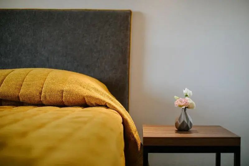 Bett mit gelber Tagesdecke, grauem Kopfteil und einem Nachttisch mit kleiner Blumenvase.