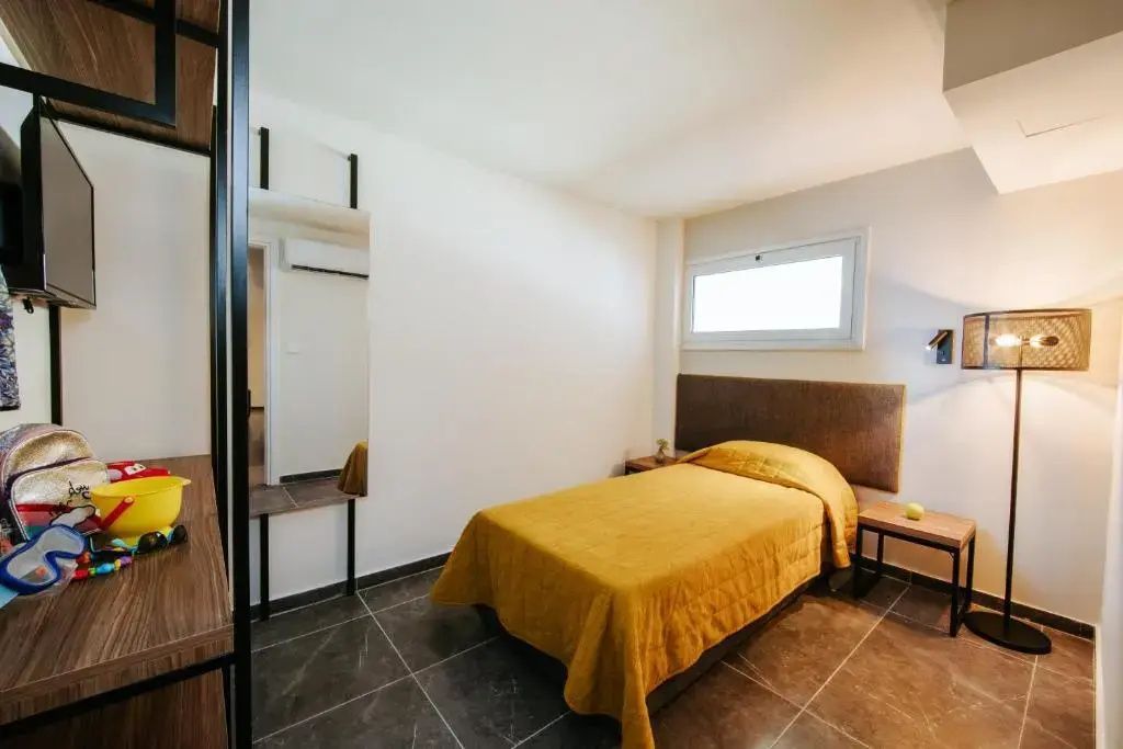 Einzelbett mit gelber Tagesdecke in einem hellen Hotelzimmer mit Fliesenboden und Spiegel.