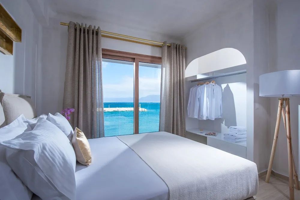 Schlafzimmer mit Blick durch das Fenster auf das Meer, gemachtem Bett und offenem Schrank.