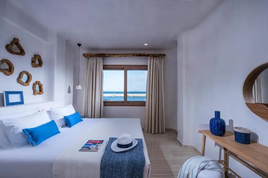 Hell eingerichtetes Schlafzimmer mit Doppelbett, blauen Kissen und Fenster mit Meerblick.