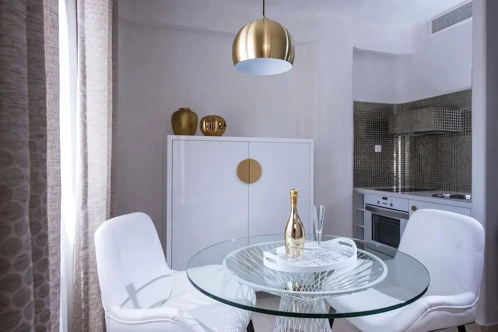 Heller Essbereich in einer Suite mit rundem Glastisch, weißem Schrank und goldener Lampe.