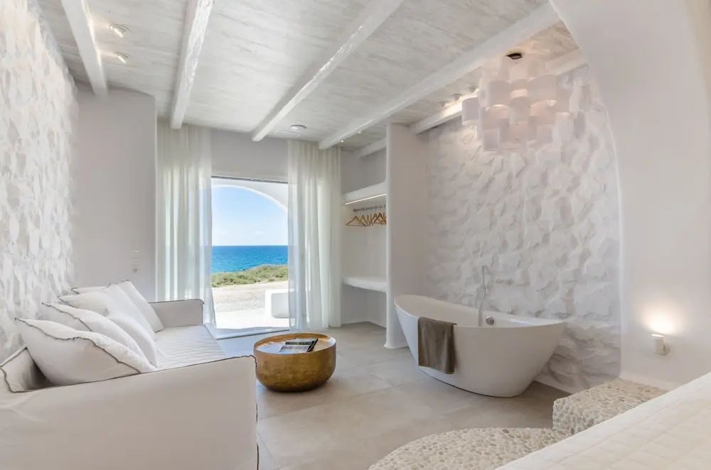 Weiß eingerichtetes Zimmer mit freistehender Badewanne, Sofa und Blick aufs Meer.