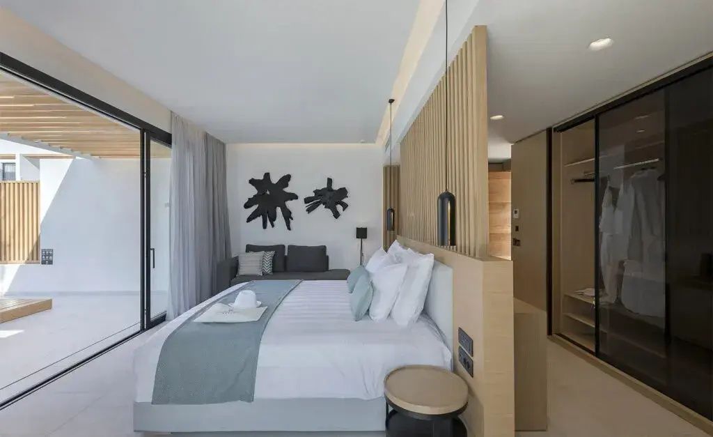 Modernes Hotelzimmer mit Doppelbett, Holztrennwand, grauer Couch und großem Balkonfenster.