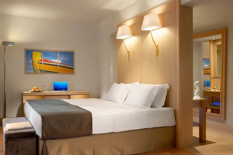 Doppelbett mit hellbraunem Bettläufer, Wandlampen und Schreibtisch in einem Hotelzimmer.
