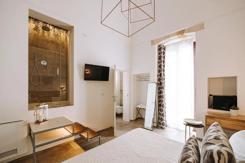 Helles Hotelzimmer mit Doppelbett, Wand-TV, bodentiefem Fenster und transparenter Duschkabine auf Podest.