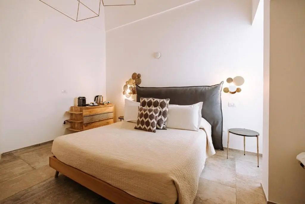 Helles Hotelzimmer mit großem Doppelbett, beiger Tagesdecke und dekorativer Beleuchtung an der Wand.
