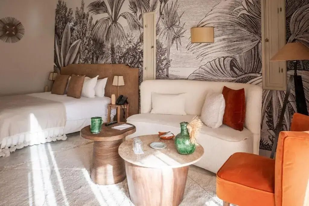 Helles Hotelzimmer mit Doppelbett, weißem Sofa, Holztischen und illustrierter Palmentapete.