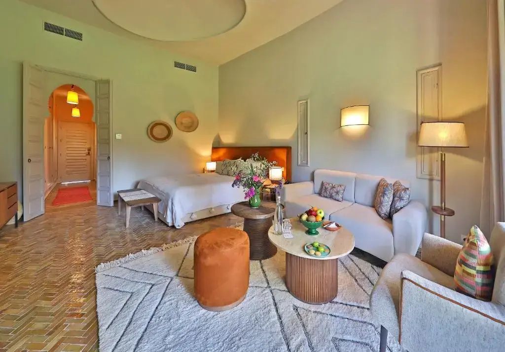 Geräumiges Hotelzimmer mit Doppelbett, hellem Sofa, Holzbeistelltischen und Teppich auf Fliesenboden.