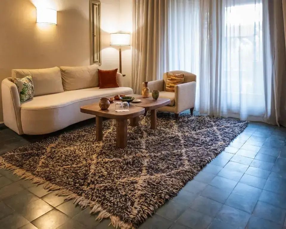 Sitzecke im Hotelzimmer mit beigem Sofa, Sessel, Holztischen und großem gemustertem Teppich.