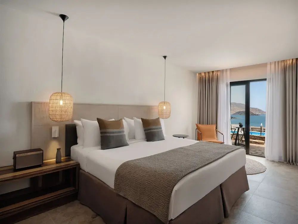 Doppelbett in hellem Hotelzimmer mit Pendelleuchten und Blick auf den Balkon und das Meer.