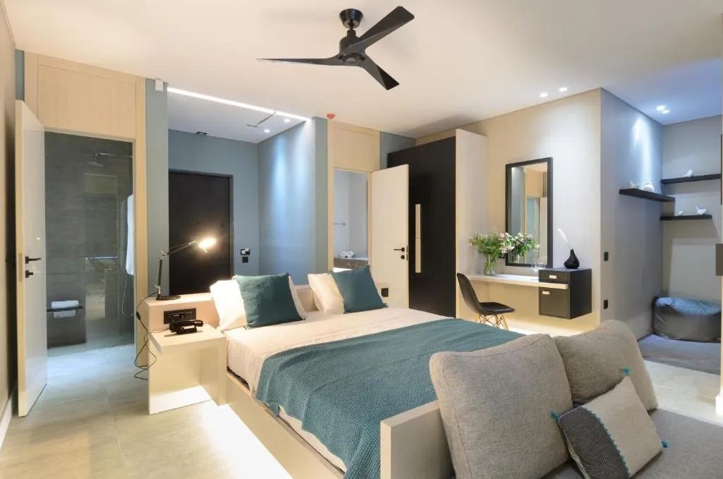 Doppelbett mit blauer Tagesdecke, Deckenventilator und kleinem Arbeitsbereich im Hotelzimmer.