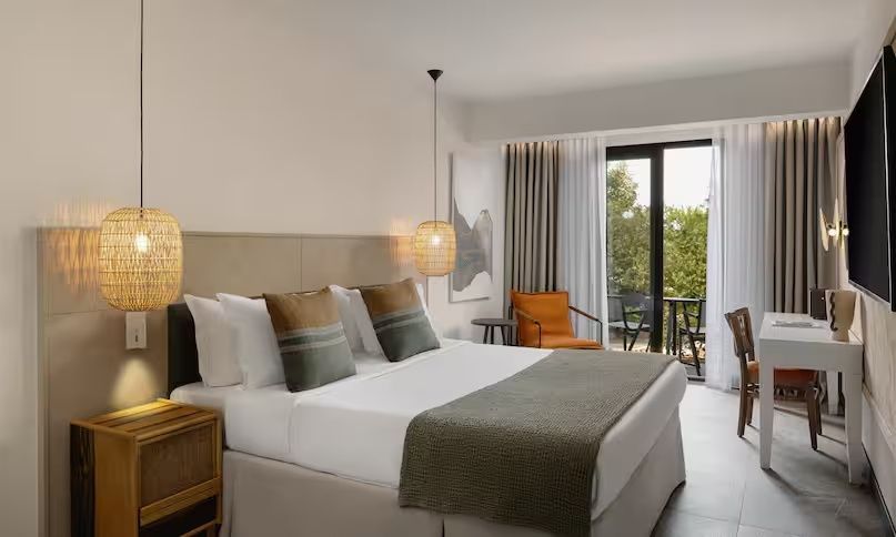 Doppelbett im Hotelzimmer mit Kissen, Sessel und Blick auf den Balkon.