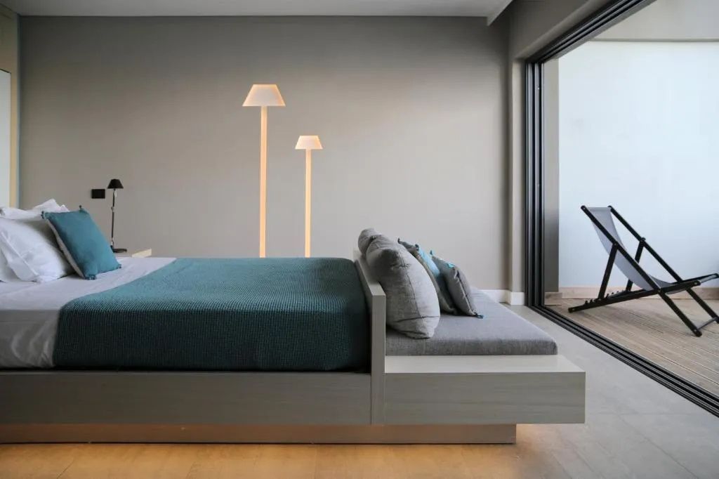 Seitliche Ansicht eines Doppelbetts mit blauer Decke und stehenden Designlampen an der Wand.